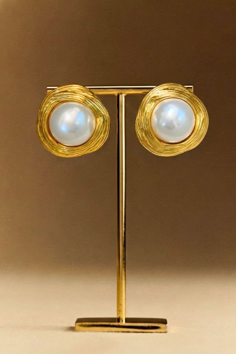 Pair of gold swirl pearl stud earrings displayed on a gold stand, showcasing round pearls framed by sculpted circular gold detailing.