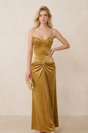 A blonde model wears a gold velvet ruched maxi dress with slim spaghetti straps and a softly draped waist, styled with gold statement earrings and a matching clutch.