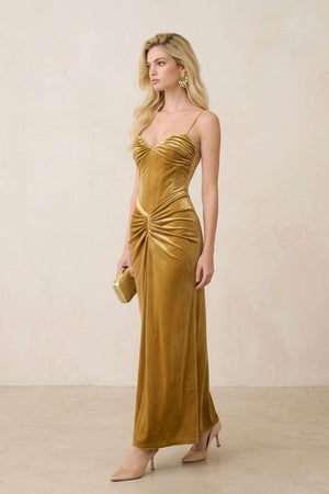 Side profile of a gold velvet ruched maxi dress worn with neutral heels, showcasing the contoured silhouette, gathered hip detail, and elegant velvet texture.
