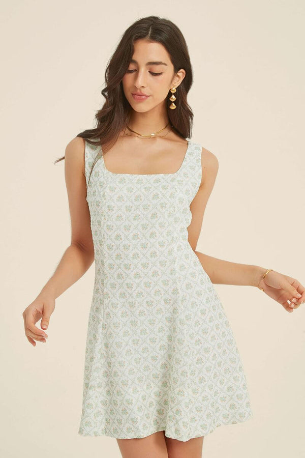 Woman wearing a light green jacquard floral mini dress with a square neckline and flared hem, styled with gold jewelry and white heels.