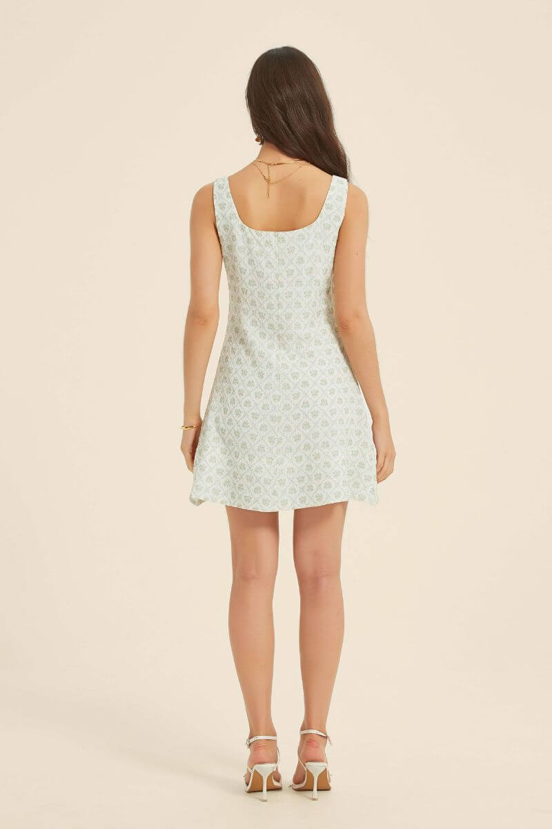 Back view of a woman in a green jacquard floral mini dress with a scoop neckline and A-line silhouette, styled with gold accessories.