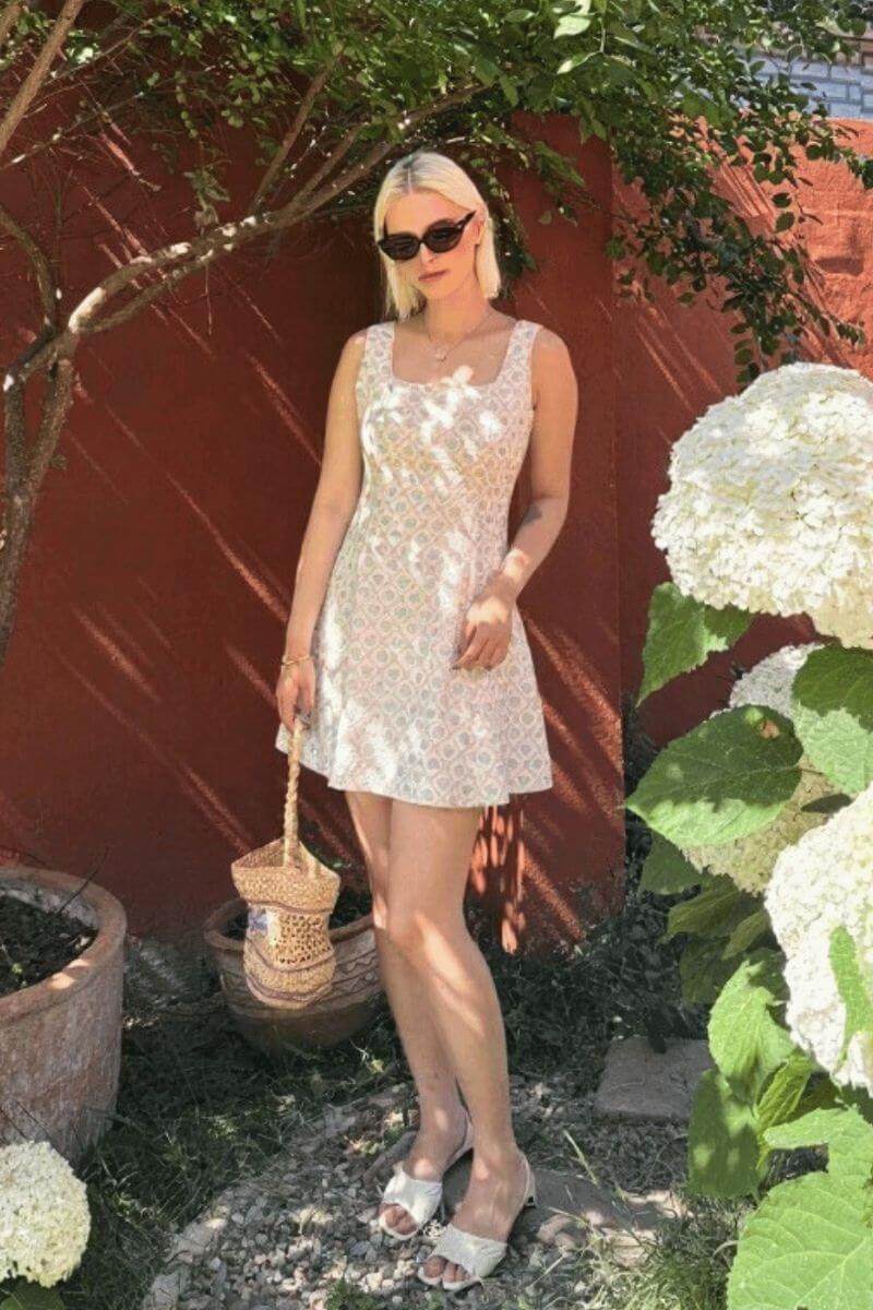 Woman wearing a green jacquard floral mini dress outdoors with a straw bag and sunglasses, standing beside white hydrangeas under sunlight.