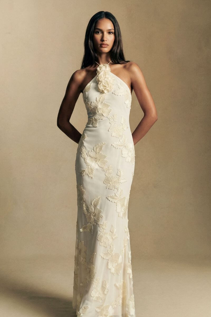Model stands front-facing in an ivory halter maxi dress featuring tonal floral embroidery and a softly fitted silhouette, photographed in warm editorial lighting.