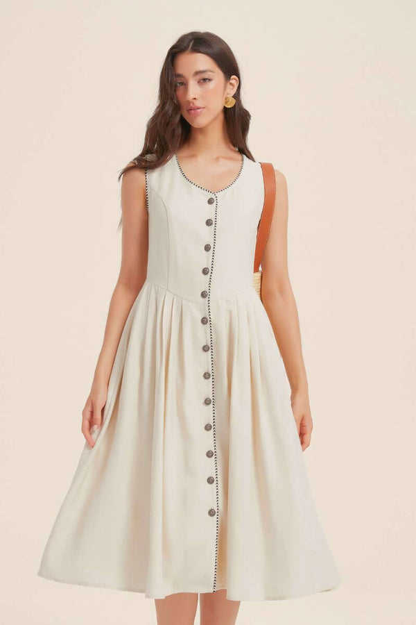 Woman wearing an ivory sleeveless midi dress with black contrast trim and front buttons, styled with gold earrings and a tan shoulder bag for an understated romantic look.