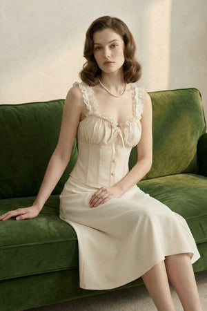 Model seated on a green velvet sofa wearing an ivory lace corset top and matching midi skirt in soft natural light.