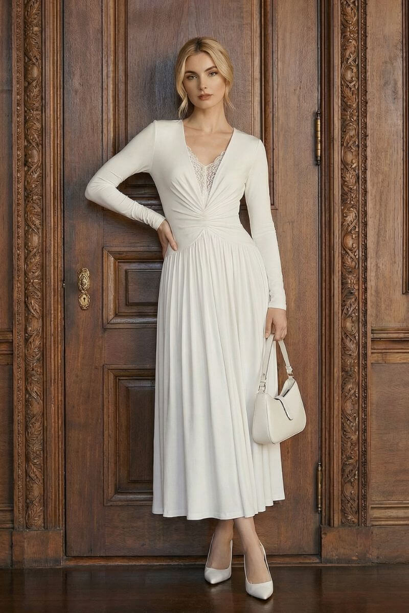 A model wears an ivory long sleeve maxi dress with a softly draped skirt and lace V-neck detail, standing in front of an ornate wooden door.