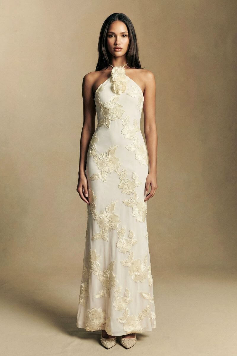 Model wears an ivory halter-neck column dress with all-over floral appliqué detailing, creating an elegant and elongated evening look.