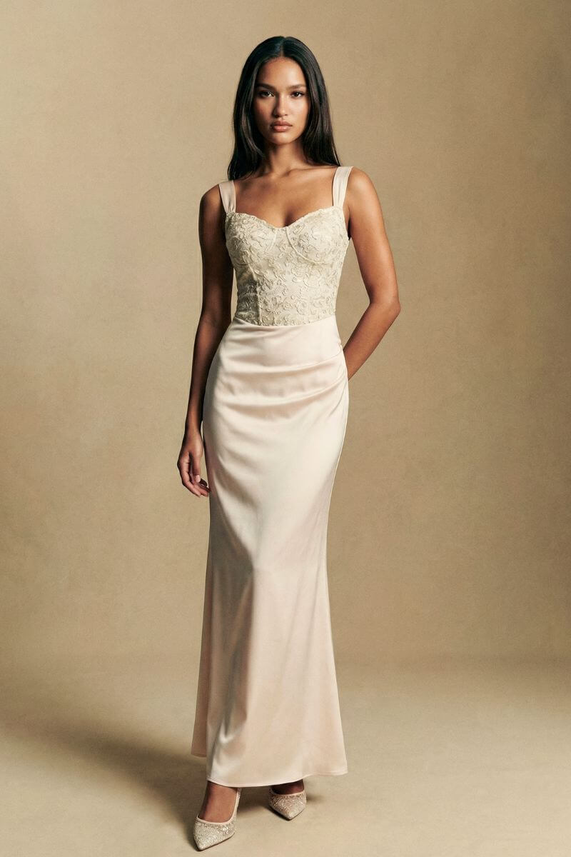 Model wearing an ivory embroidered satin maxi dress with a fitted bodice, wide straps, and a smooth column skirt, styled in a warm neutral studio setting.