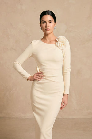 A model wearing an ivory long-sleeve midi dress with a sculpted silhouette and soft floral appliqué detail on the shoulder, styled against a warm neutral studio backdrop.