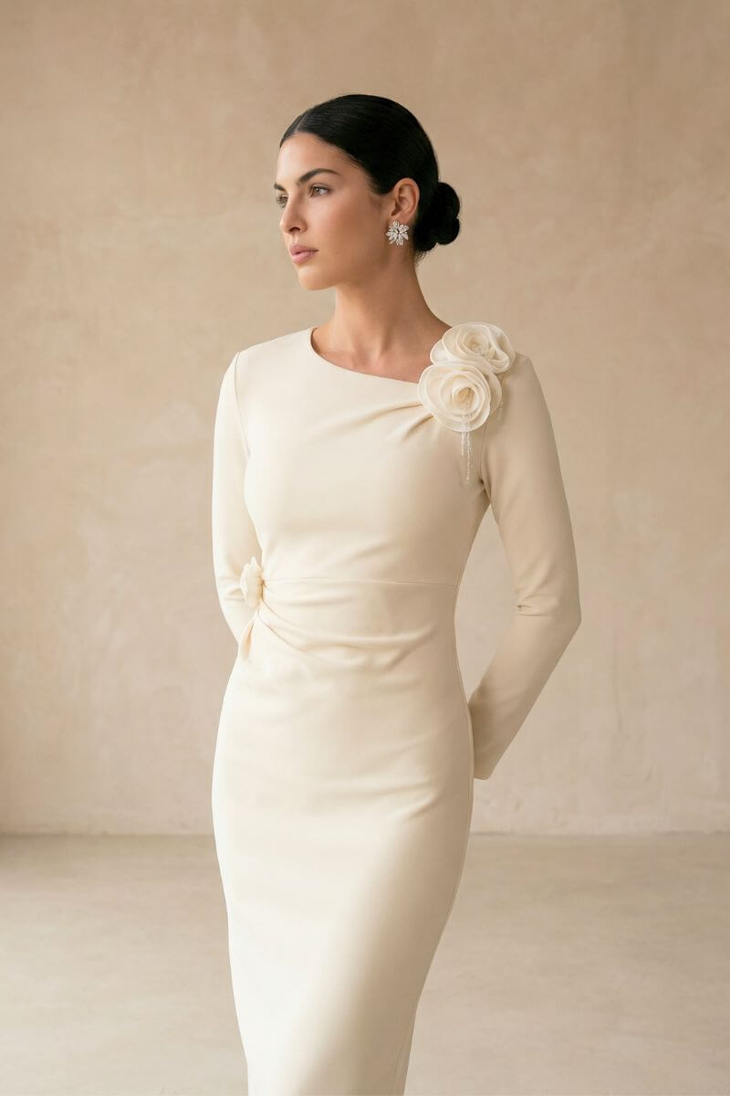 A side-angle view of a model wearing an ivory long-sleeve midi dress with a floral shoulder accent and elegant, body-skimming fit in a neutral studio setting.