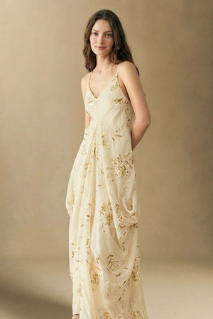 Model posing in an ivory maxi dress with gold sequin accents, thin straps, and a gently gathered waist, creating an elegant, feminine silhouette.