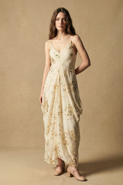Model wearing an ivory maxi dress with subtle gold sequin floral motifs, a V-neckline, and fluid drape, styled with neutral heels in a softly lit studio setting.