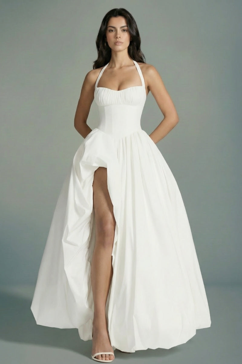 Model wears an ivory halter corset maxi dress with a softly gathered bodice, cinched waist, and dramatic front slit, styled with minimal heels against a clean studio background.