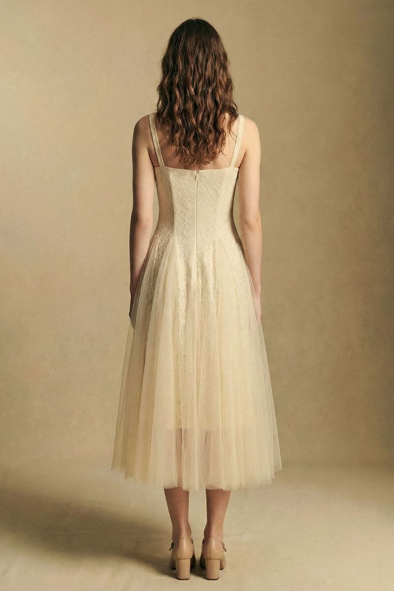 Model shown from the back wearing an ivory lace midi dress with delicate shoulder straps and a softly layered tulle skirt, styled with neutral heels against a warm studio background.