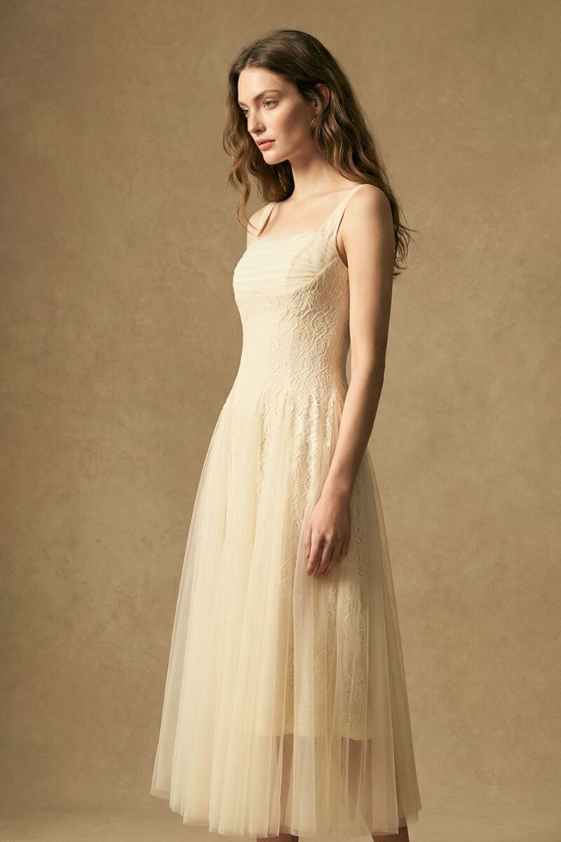 Side view of a model in an ivory lace midi dress highlighting the fitted waist, delicate lace texture, and flowing tulle skirt, captured in warm natural studio light.