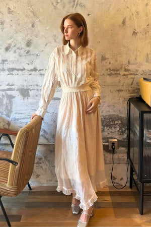 Model wearing a vintage-inspired ivory long sleeve maxi dress featuring pleated bodice, lace stripes, and soft sheen fabric, standing beside a textured wall.