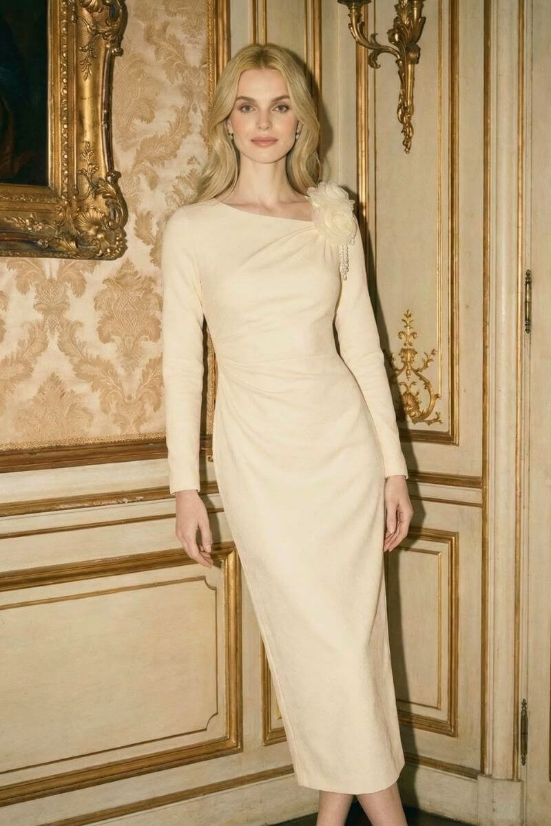 Model posing in an ivory long-sleeve midi dress with a sculpted silhouette and floral shoulder embellishment inside a classic ornate room.