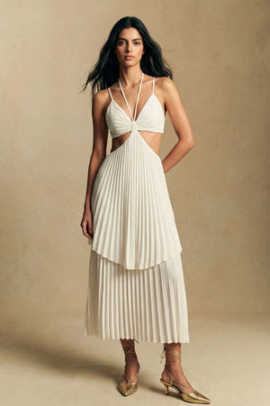 Model wears an ivory pleated midi dress with delicate cut-out waist detailing and a layered skirt, styled with gold lace-up heels against a warm neutral studio backdrop.