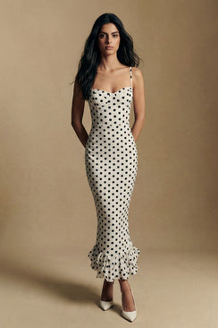 A model faces the camera in an ivory polka dot spaghetti-strap midi dress with a smooth fitted bodice and softly flared ruffle hem, styled in a minimal studio setting.