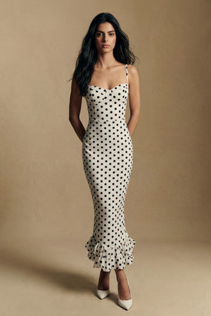 A model faces the camera in an ivory polka dot spaghetti-strap midi dress with a smooth fitted bodice and softly flared ruffle hem, styled in a minimal studio setting.