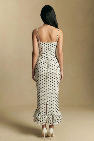 A back view of a model wearing an ivory polka dot midi dress with delicate straps, a lace-up bodice detail, and a softly ruffled hem in a clean studio environment.