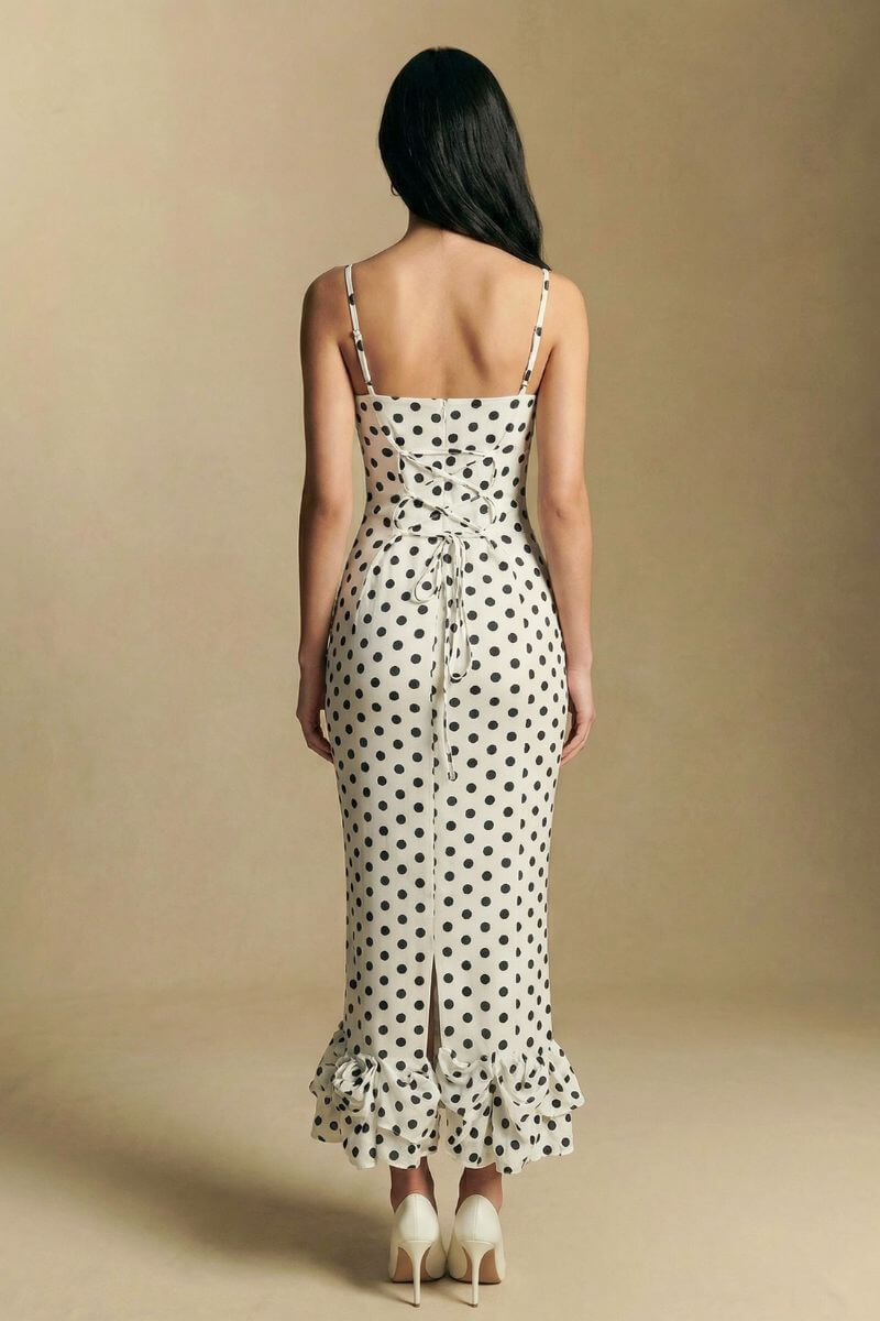A back view of a model wearing an ivory polka dot midi dress with delicate straps, a lace-up bodice detail, and a softly ruffled hem in a clean studio environment.