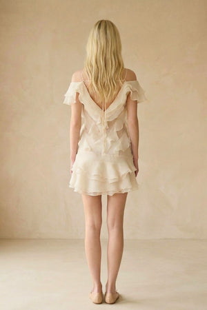 The back view of an ivory off-shoulder mini dress reveals soft ruffled layers and a lightweight chiffon finish flowing gently over the skirt.