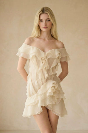 A blonde model faces forward in an ivory off-shoulder mini dress featuring cascading chiffon ruffles and a lightweight, romantic silhouette against a neutral studio backdrop.