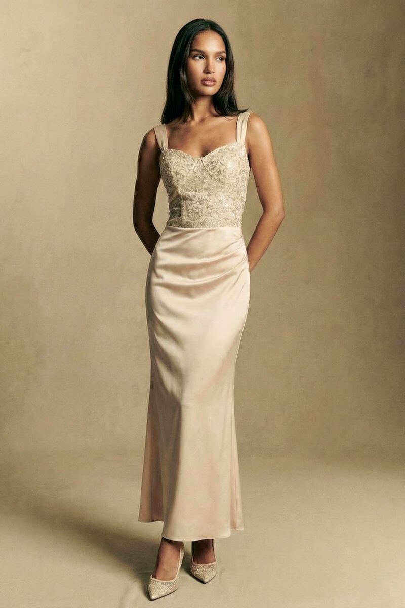 Editorial-style image of a model wearing an ivory satin maxi dress with an embroidered fitted bodice and floor-length skirt, photographed in soft neutral lighting.