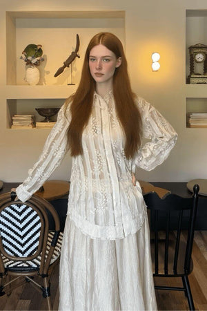 Model wearing an ivory sheer embroidered lace blouse with pleated stripe texture and floral detailing on long sleeves.