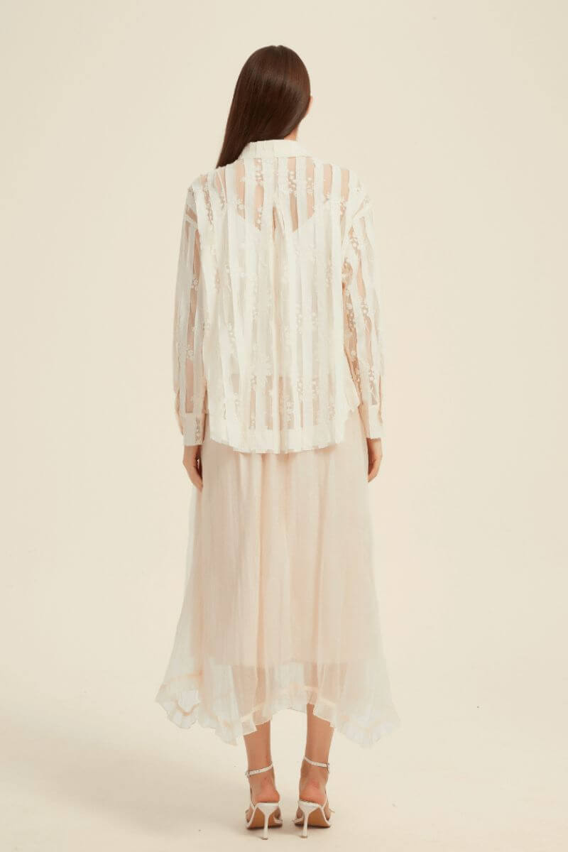 Woman wearing an ivory sheer striped lace blouse paired with a blush pink flowy midi skirt and white strappy heels, photographed against a neutral background.