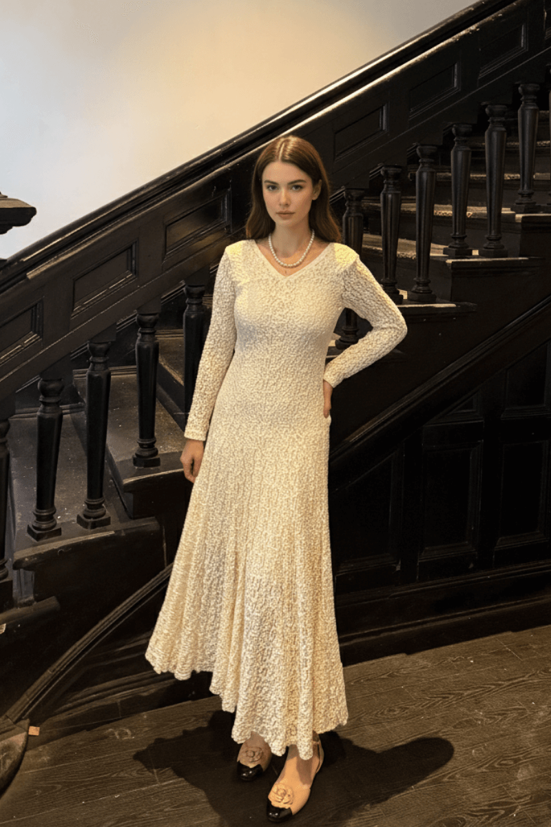Model standing by a staircase in an ivory textured maxi dress with long sleeves, accessorized with pearl jewelry and nude heels.