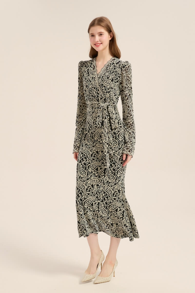 A woman in a black and beige embroidered wrap midi dress poses in profile, showing its fitted waist, flared hemline, and romantic lace detailing.