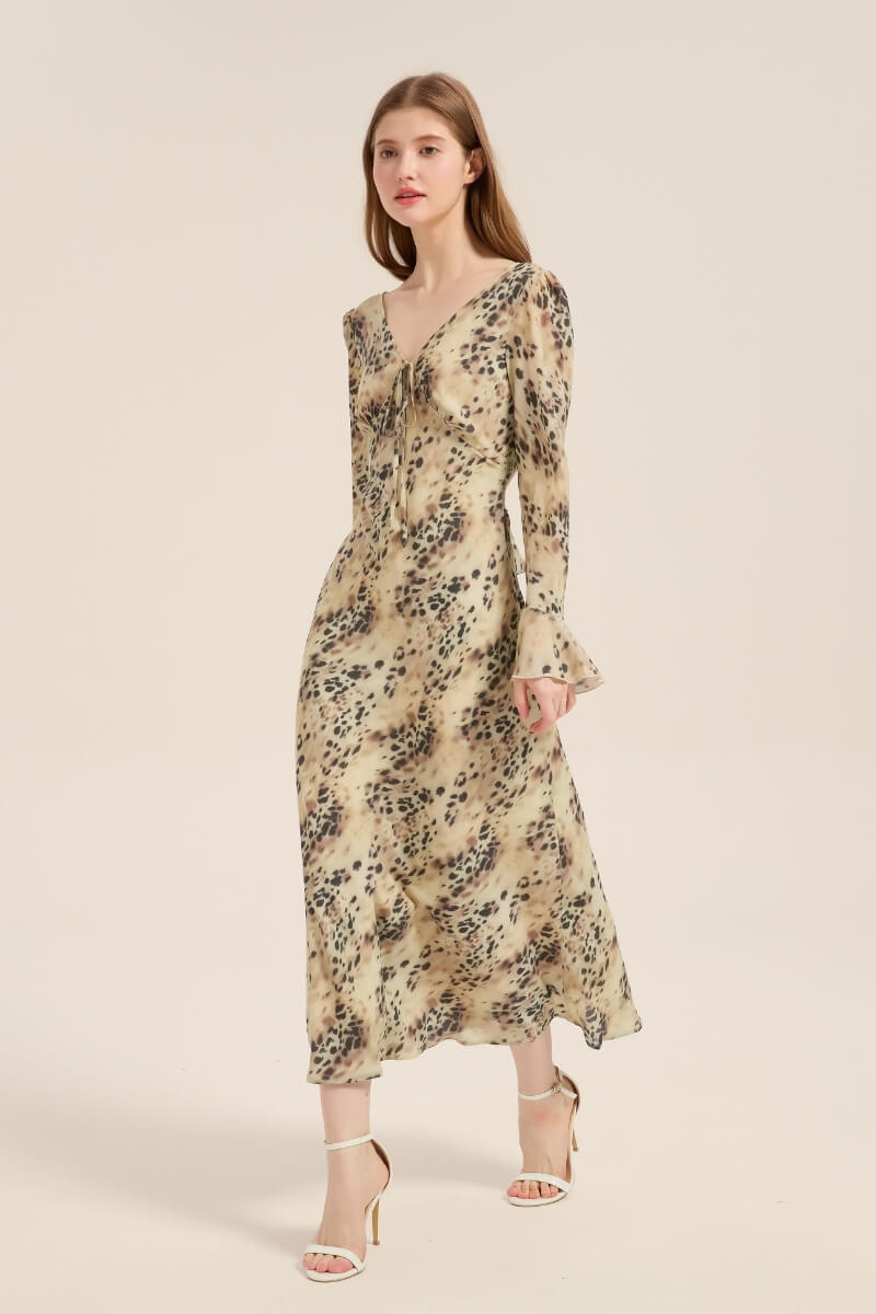 Model walking in a beige leopard print satin midi dress with a V-neckline, tie front, and flared cuffs, styled with white heels on a neutral backdrop.