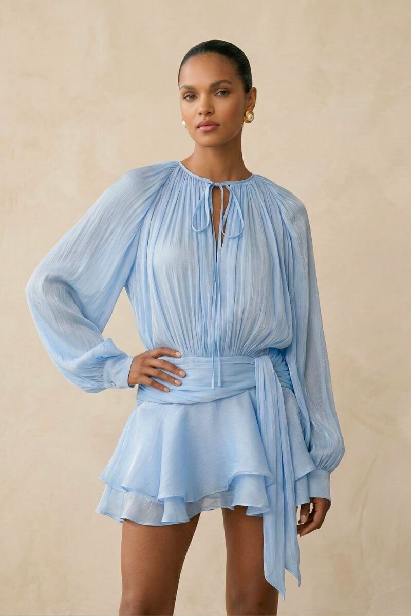 Model posed slightly angled in a light blue chiffon mini dress featuring airy pleats, a soft tie neckline, gathered waist, and flowing ruffle hem for a feminine silhouette.