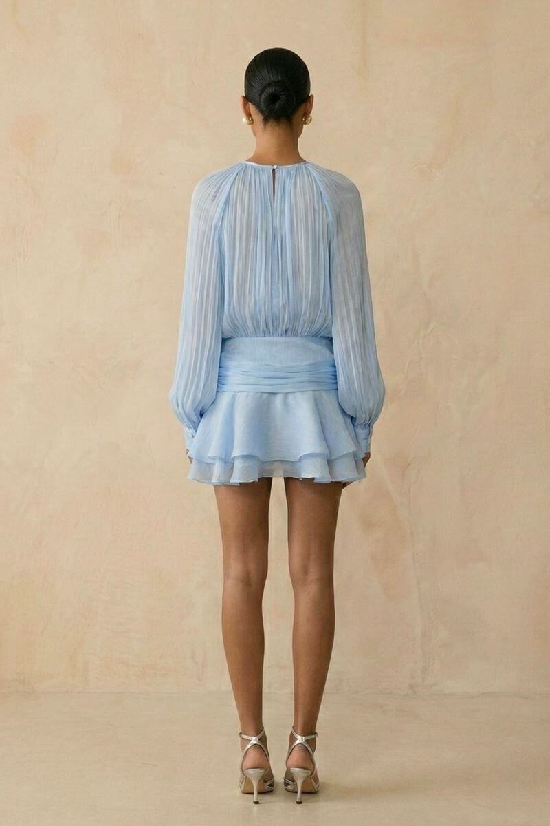 Model shown from the back wearing a light blue chiffon mini dress with soft pleated long sleeves, a gathered waist sash, and a layered ruffle hem against a warm neutral studio background.