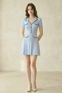 A woman wearing a light blue knit A-line mini dress featuring pearl buttons, pleated texture, and contrast black piping, photographed full length from the front.