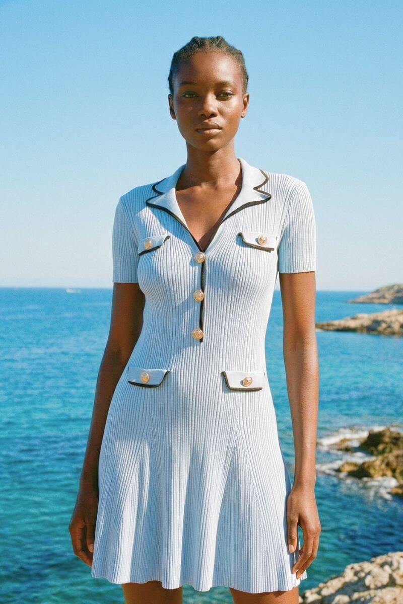 Close-up of a woman in a light blue knit mini dress near the ocean, showing pearl buttons and contrast trim.