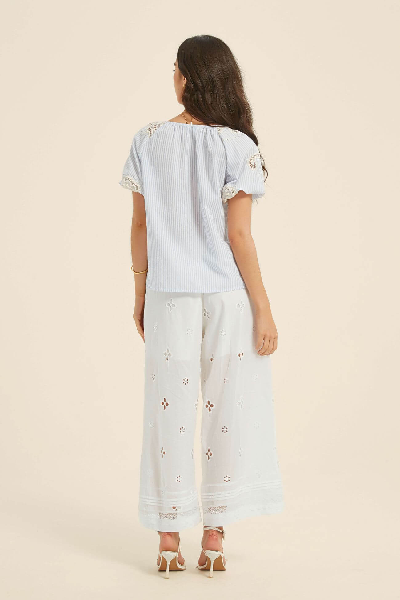 Full-length view of a woman wearing a light blue striped boho blouse with lace inserts and tassel ties, paired with white wide-leg embroidered pants and strappy heels for a breezy, coastal look.
