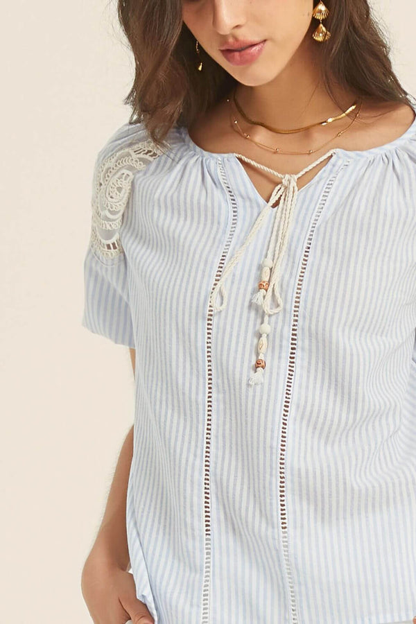 Close-up of a light blue striped blouse with crochet lace shoulders and tasseled drawstring neckline, styled with gold layered necklaces and earrings.