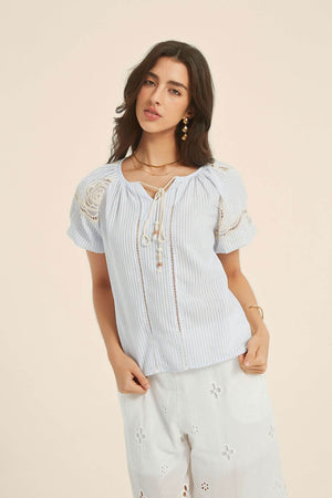 Front view of a woman wearing a light blue striped blouse with lace shoulder accents and tie-front tassels, paired with white embroidered trousers for an airy resort look.