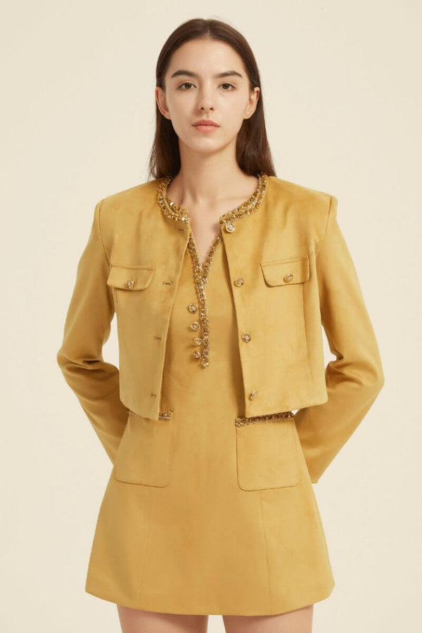 Model wearing a mustard yellow two-piece outfit featuring a cropped jacket and sleeveless mini dress, both accented with gold beaded trim and buttons — sophisticated fall ensemble.