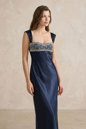 A three-quarter view of a navy satin maxi dress highlighting the beaded bust panel, sculpted waist, and elegant drape of the skirt.