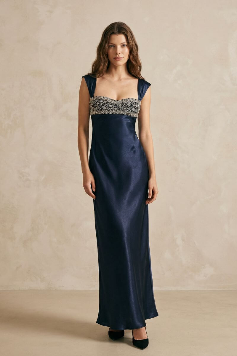 A full-length front view of a navy satin maxi dress with wide straps, ornate beaded bust embroidery, and a softly flowing skirt in a minimalist studio setting.