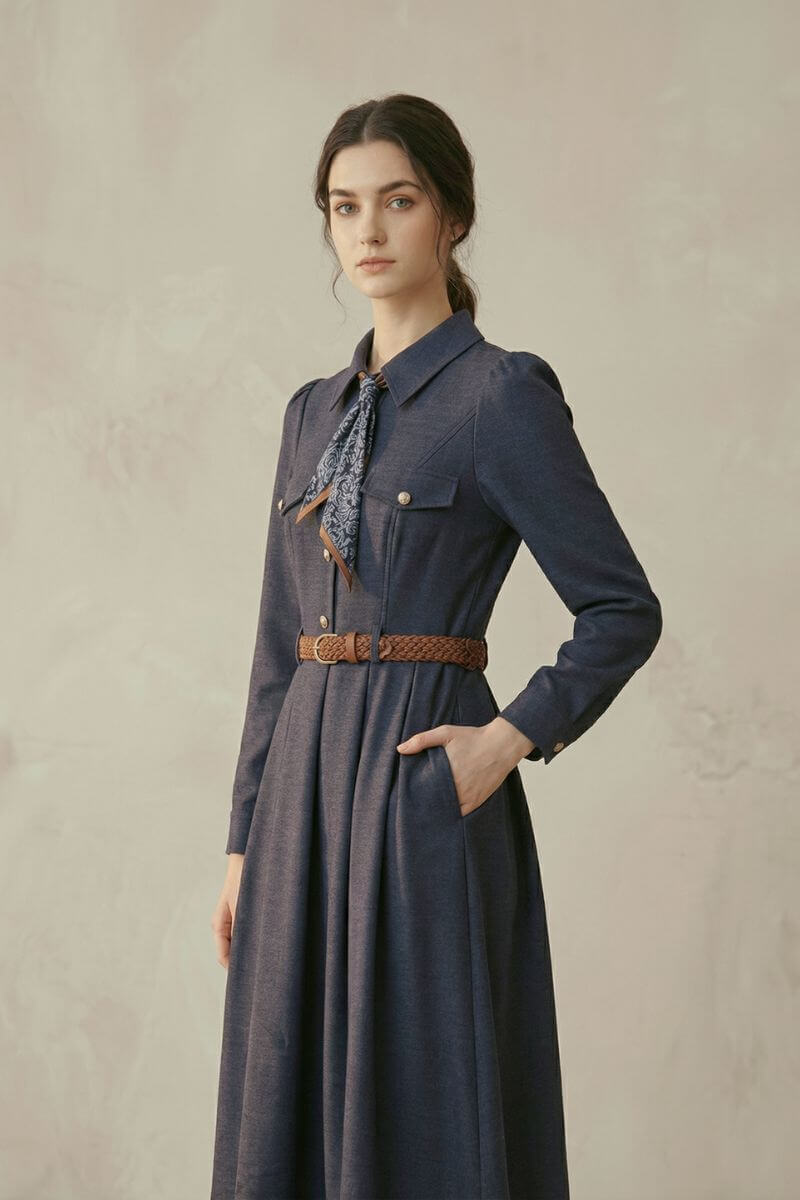 A woman in a navy collared midi dress featuring long sleeves, gold-tone buttons, a printed neck scarf, and a brown braided belt, shown from an angled front pose.