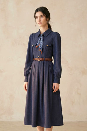 A woman wearing a navy long-sleeve collared midi dress with gold-tone buttons, a printed neck scarf, and a brown woven belt, photographed from a soft side angle.