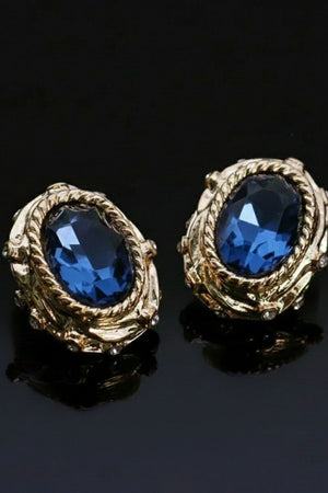 Angled close-up of gold stud earrings with oval navy crystal centers and sculptural rope-textured settings, photographed on a glossy black surface.