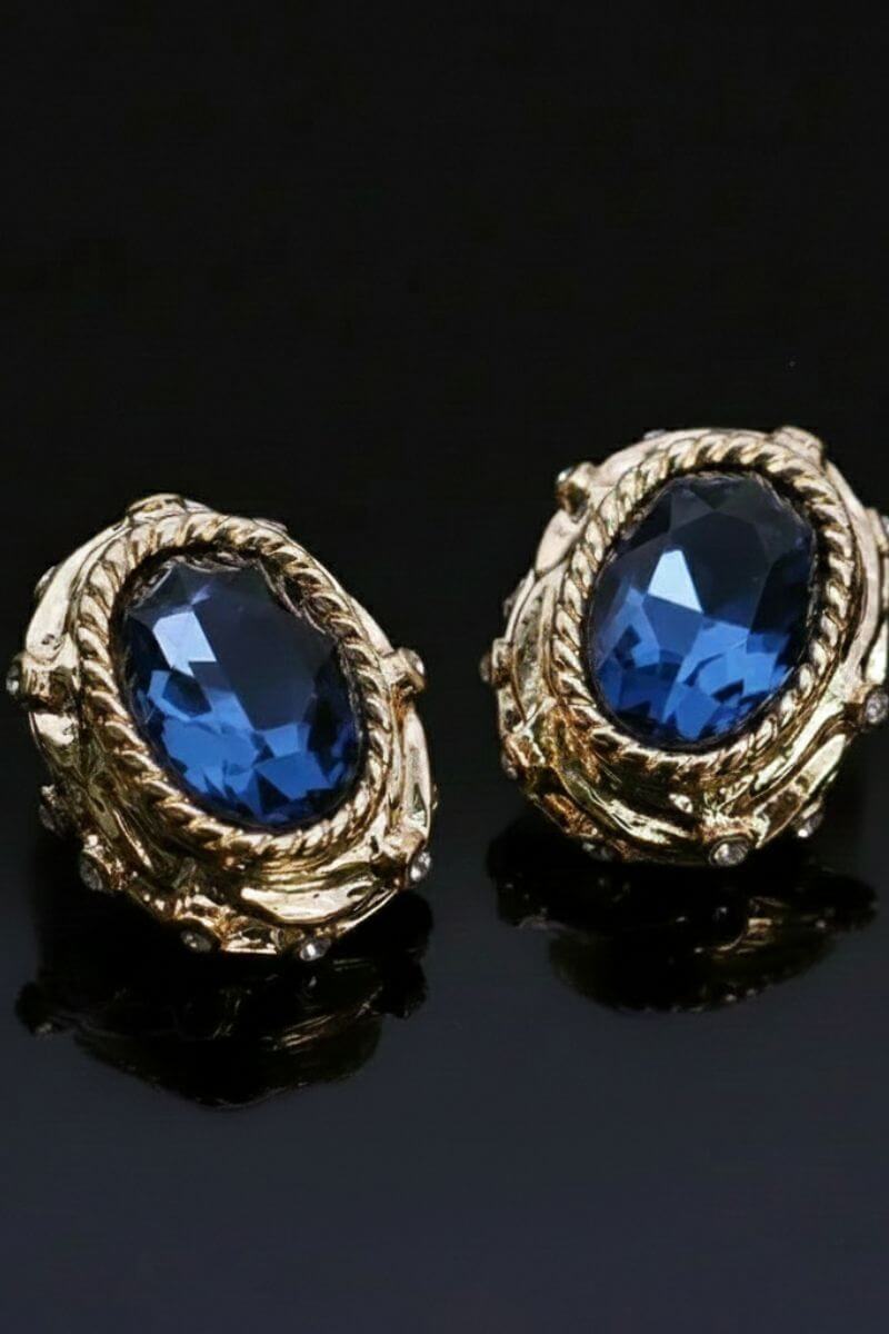 Angled close-up of gold stud earrings with oval navy crystal centers and sculptural rope-textured settings, photographed on a glossy black surface.