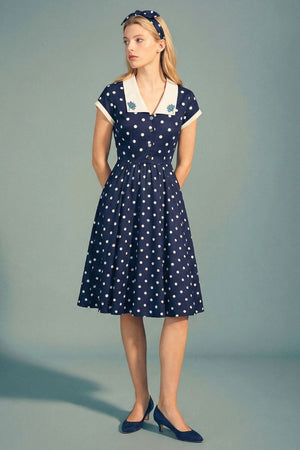 A model stands in a navy polka dot midi dress featuring an ivory contrast collar, short sleeves, and a gently flared skirt, paired with navy pumps in a minimalist studio setting.