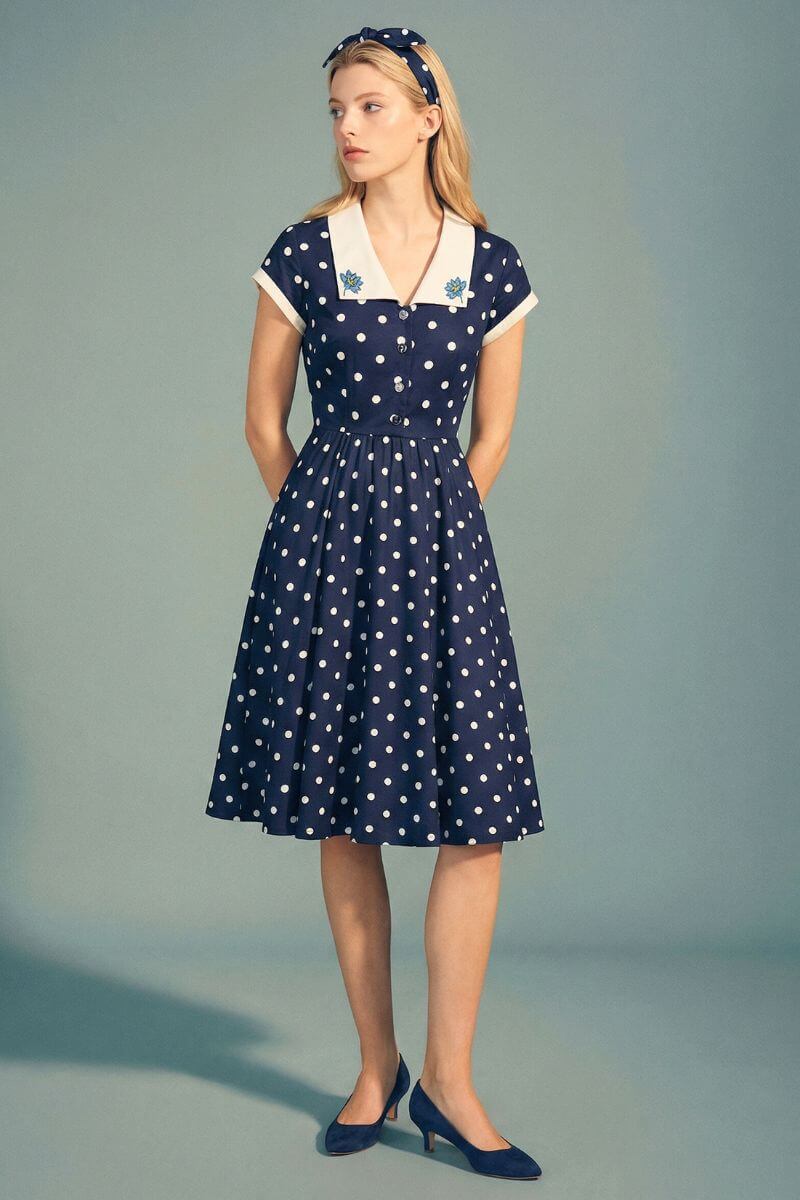 A model stands in a navy polka dot midi dress featuring an ivory contrast collar, short sleeves, and a gently flared skirt, paired with navy pumps in a minimalist studio setting.
